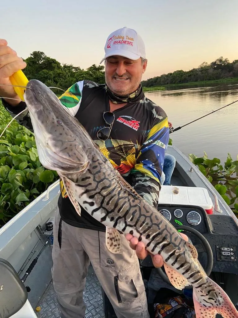 Pescador sorridente segurando um grande surubim no barco ao entardecer, com camiseta de pesca personalizada