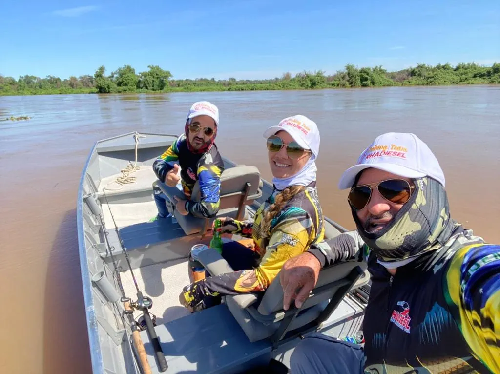 Três amigos pescando juntos num barco no rio, vestindo camisetas de pesca personalizadas e bonés da equipe