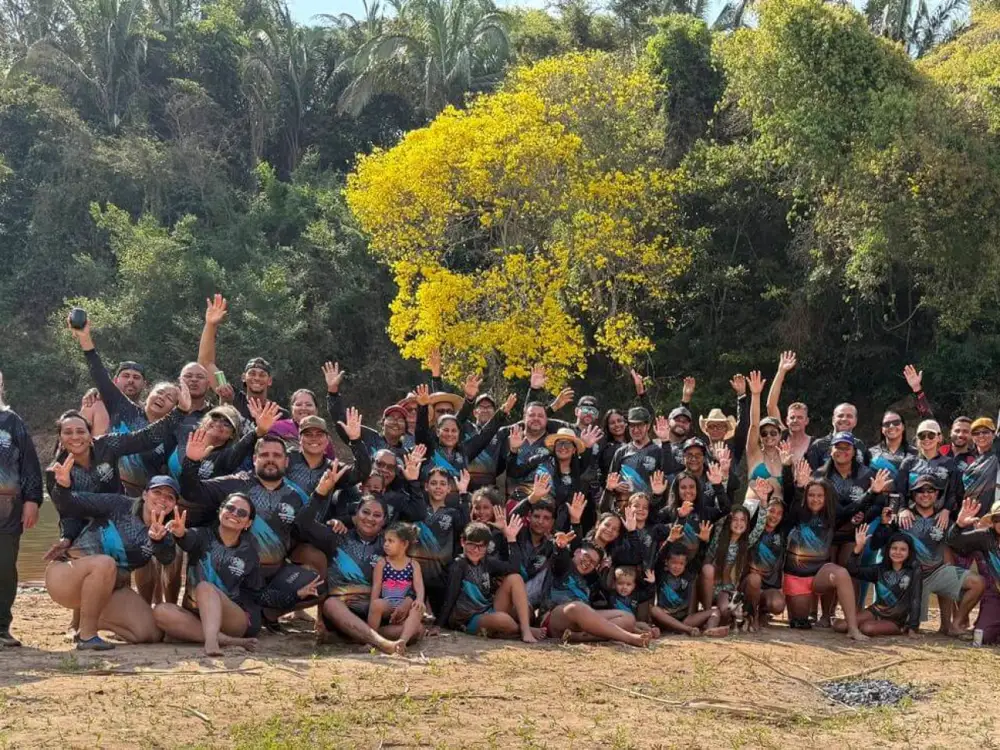Grande grupo de pescadores e famílias acenando à beira do rio, todos com camisetas personalizadas iguais