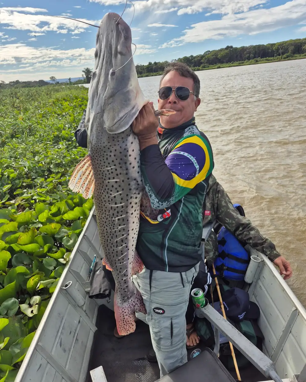 Pescador no rio exibindo um grande peixe ao lado do barco, com camiseta de pesca personalizada