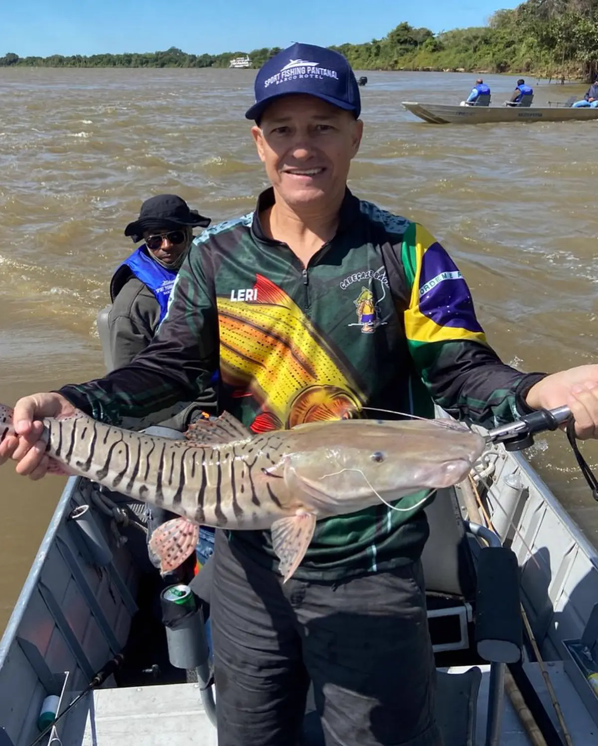 Pescador em barco no rio segurando um peixe, vestindo camiseta de pesca personalizada com a bandeira do Brasil